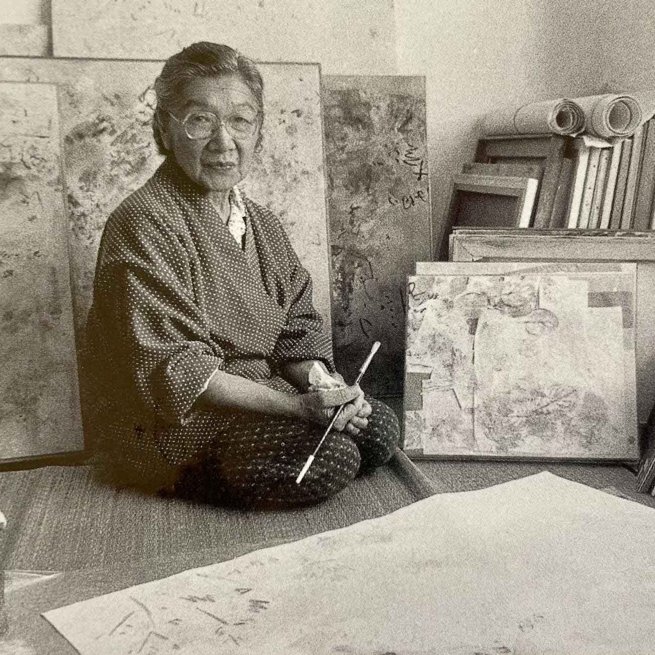 A black and white photograph of an elderly Hisako Hibi sitting on the floor of her workshop surrounded by her paintings, holding a paint brush while looking at the camera.